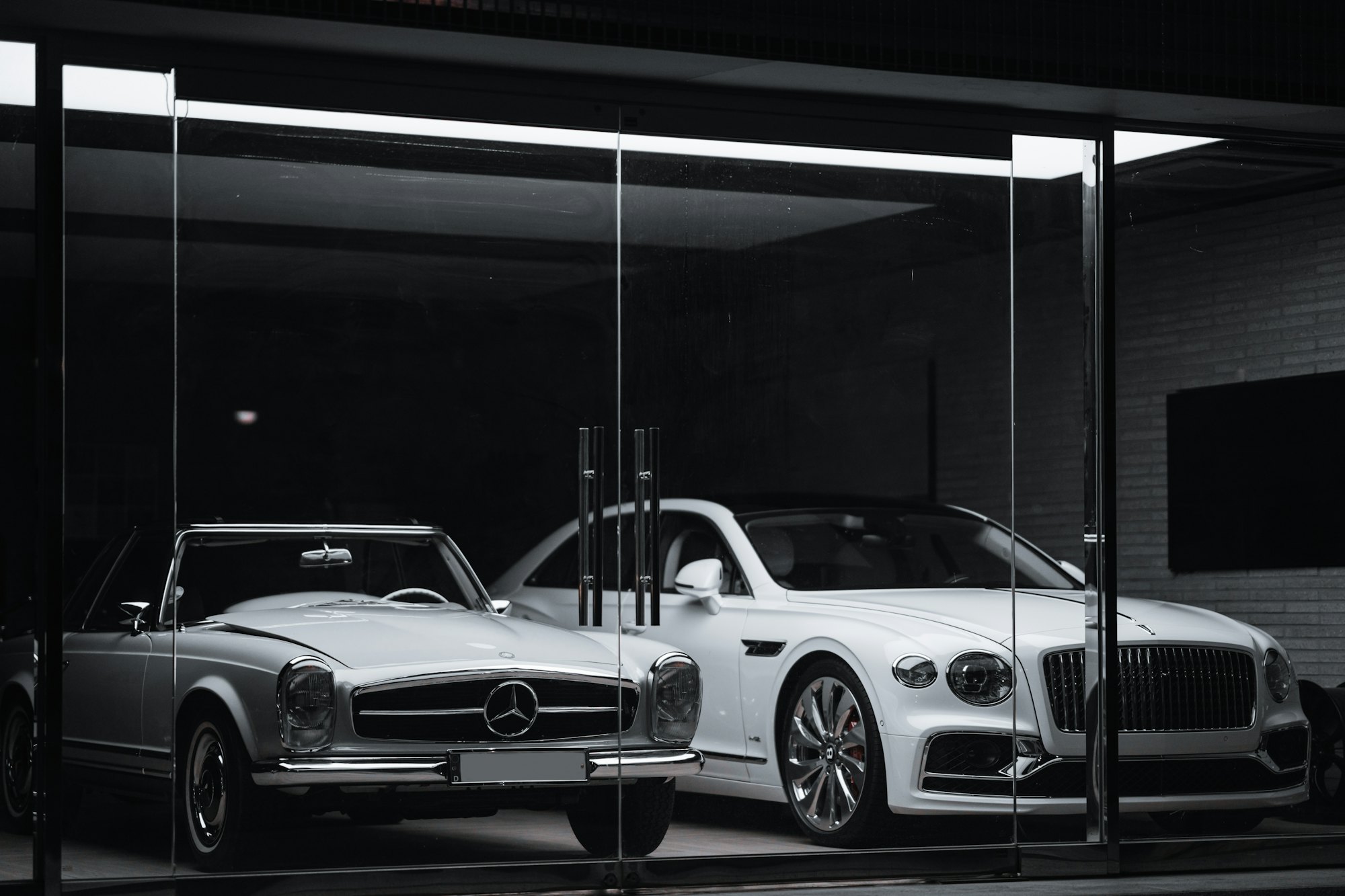 Cam vitrinli showroomda yan yana duran klasik bir roadster ve beyaz bir lüks sedan, siyah beyaz