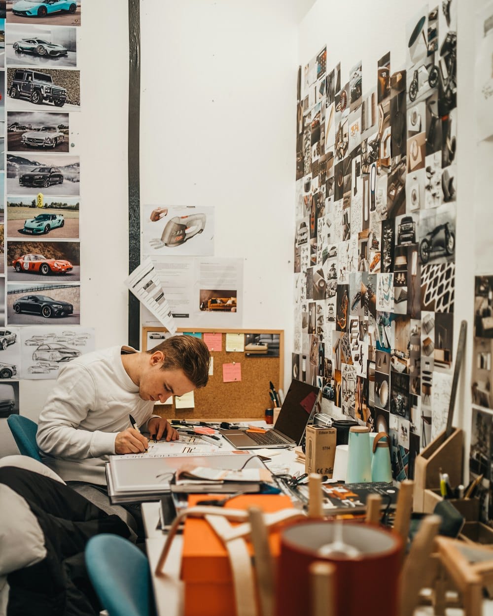A designer drawing at a desk in a studio whose wall is covered in photos and sketches